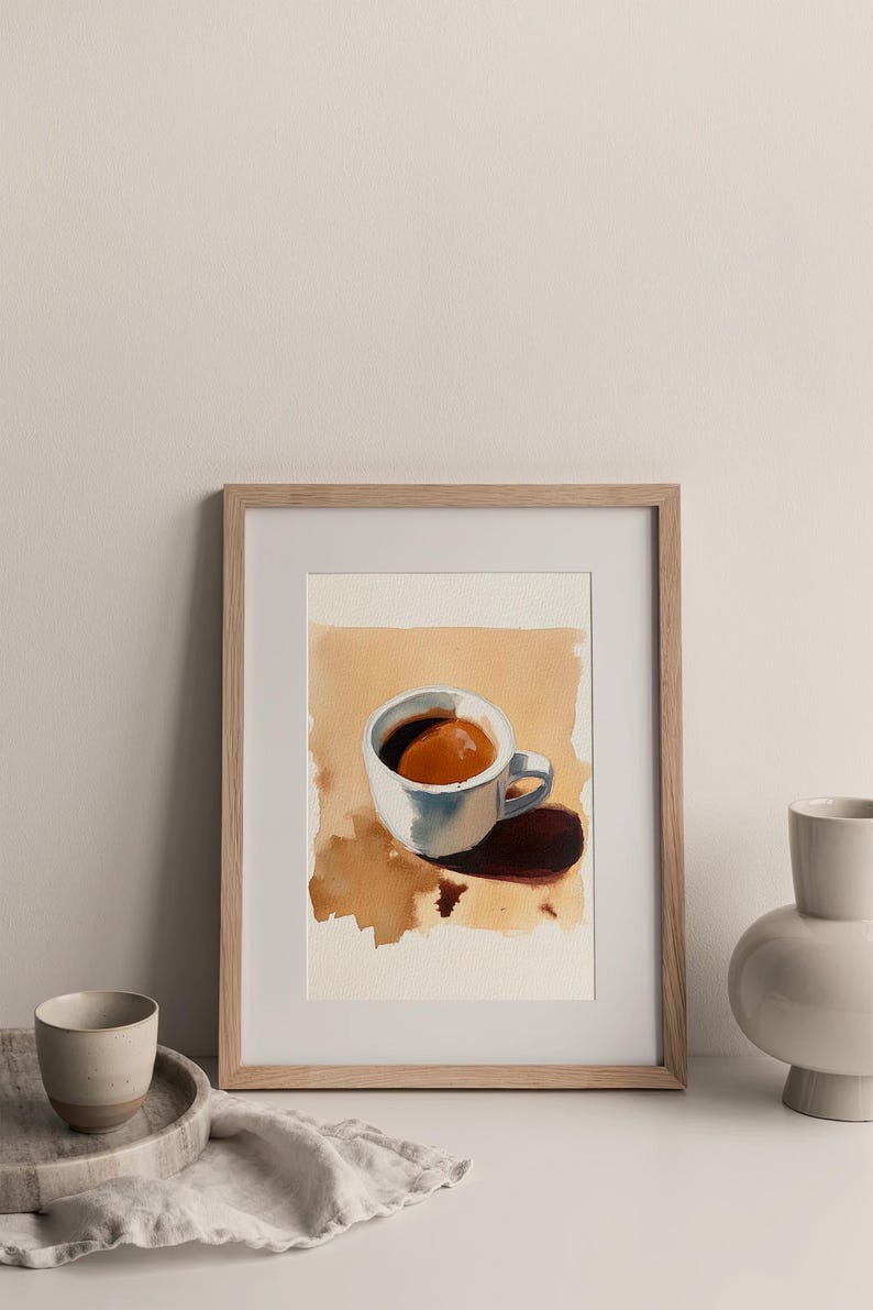 Puede incluir: Una pintura de acuarela enmarcada de una taza de caf&eacute; blanca con un l&iacute;quido marr&oacute;n en su interior. La taza est&aacute; sentada sobre una superficie marr&oacute;n claro con una sombra debajo. La pintura est&aacute; en un marco de madera claro.