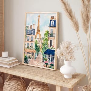 May include: A watercolour painting of a Parisian street scene with the Eiffel Tower in the background. The painting features buildings with balconies, trees, and a cafe with a blue awning. The painting is framed in a light wood frame.