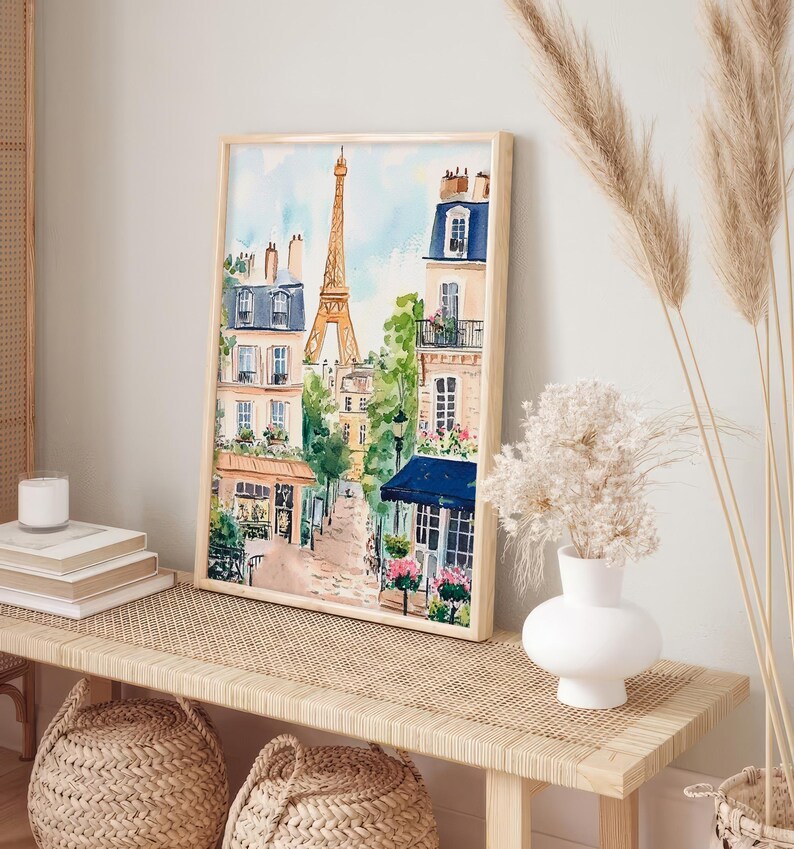 May include: A watercolour painting of a Parisian street scene with the Eiffel Tower in the background. The painting features buildings with balconies, trees, and a cafe with a blue awning. The painting is framed in a light wood frame.