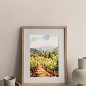 May include: A framed watercolor painting of a vineyard landscape. The painting depicts a winding path through rows of grapevines, with a small village in the distance. The sky is a light blue with fluffy white clouds.