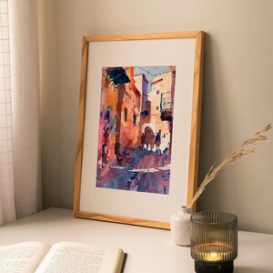 May include: Watercolor painting of a European street scene in a wooden frame. The artwork features buildings in shades of orange, pink, and blue, with figures in the street. The painting is displayed on a white surface with a book, vase, and candle.