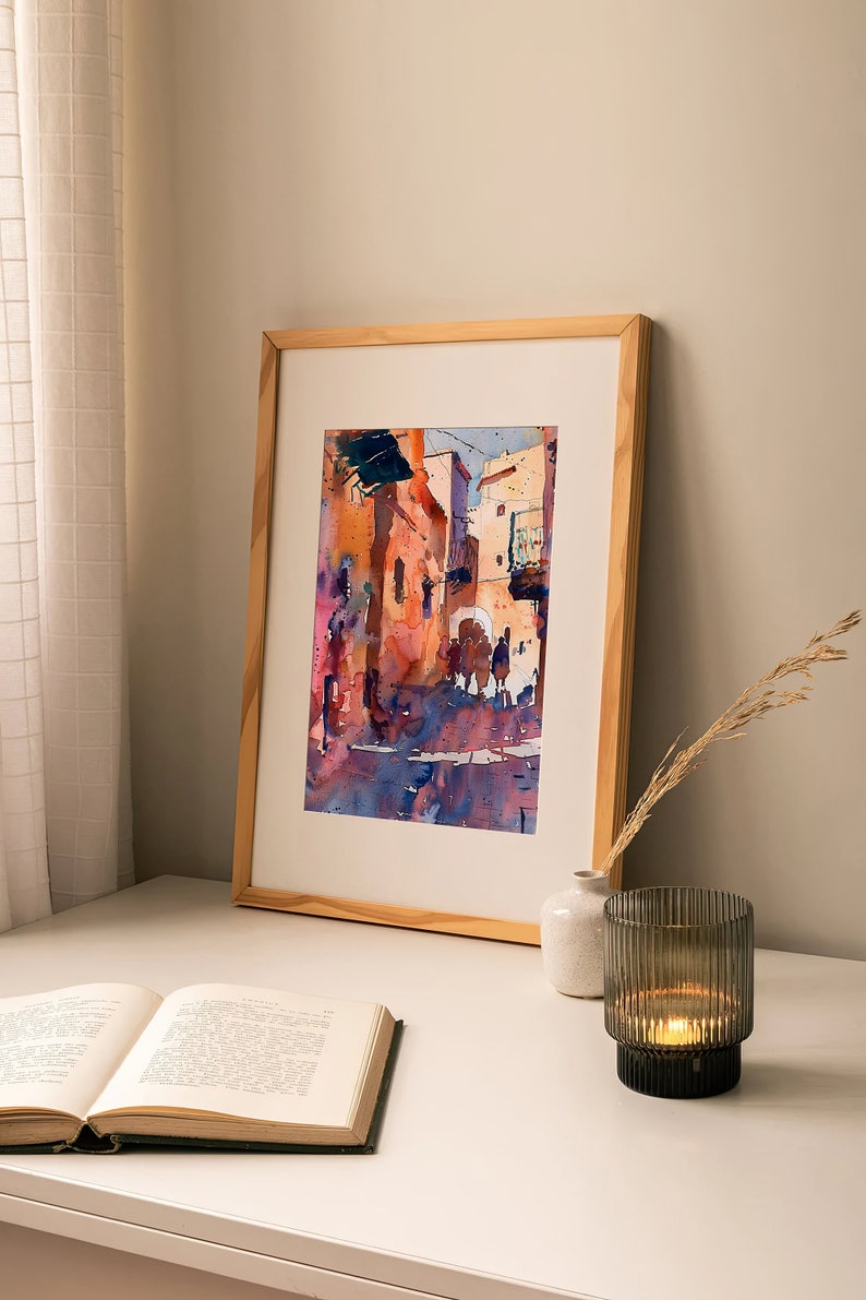 May include: Watercolor painting of a European street scene in a wooden frame. The artwork features buildings in shades of orange, pink, and blue, with figures in the street. The painting is displayed on a white surface with a book, vase, and candle.