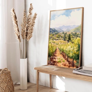 May include: Watercolor painting of a vineyard landscape with rolling hills, green vines, and a blue sky with white clouds. The painting is framed in a light wood frame and is hanging on a white wall.