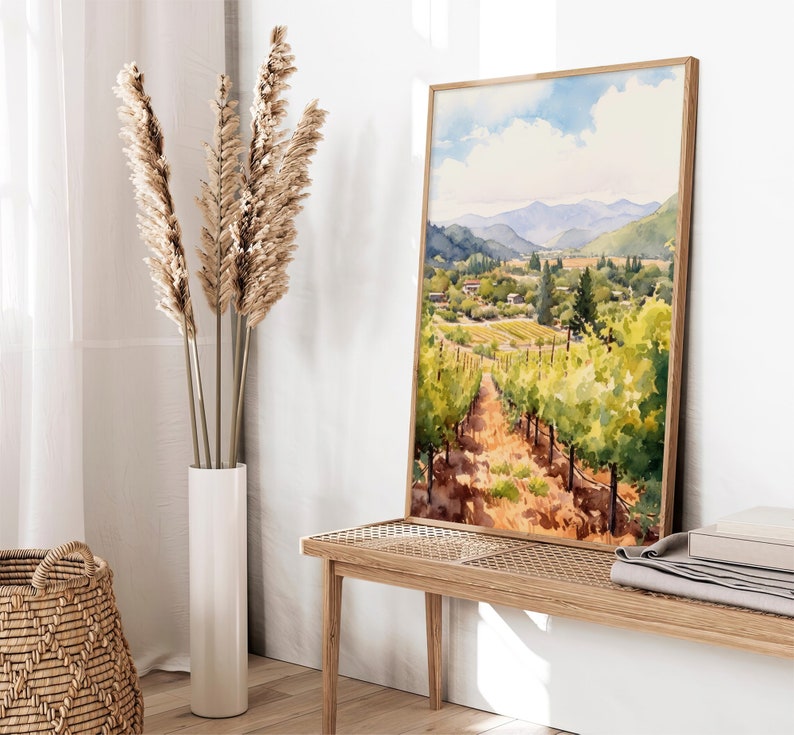 May include: Watercolor painting of a vineyard landscape with rolling hills, green vines, and a blue sky with white clouds. The painting is framed in a light wood frame and is hanging on a white wall.