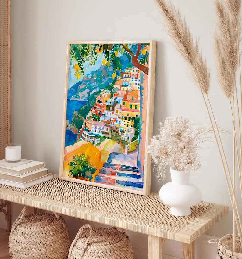 May include: Watercolour painting depicting a coastal Italian village with vibrant buildings, a blue sea, and a lemon tree. The artwork is framed in a light wood frame and displayed on a woven bench with decorative baskets.