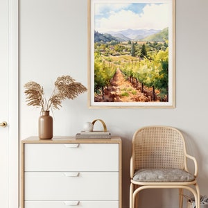 May include: A watercolor painting of a vineyard landscape with rolling hills, green vines, and a dirt path leading through the center. The painting is framed in a light wood frame and hangs on a white wall.
