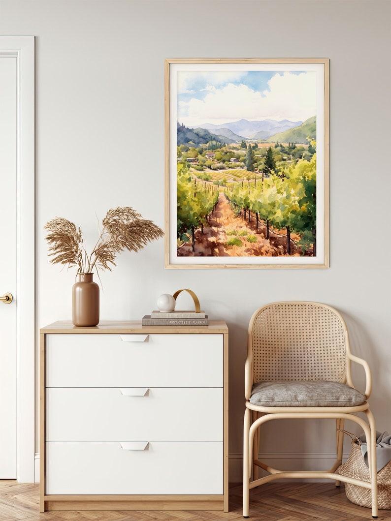 May include: A watercolor painting of a vineyard landscape with rolling hills, green vines, and a dirt path leading through the center. The painting is framed in a light wood frame and hangs on a white wall.