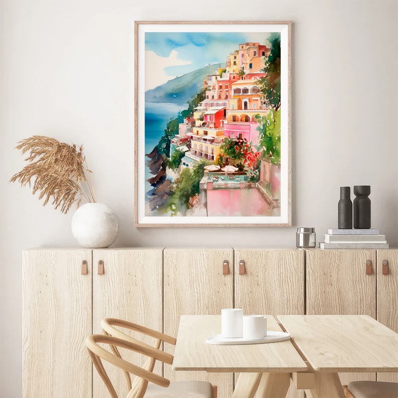 May include: Watercolour painting of colourful buildings on a hillside overlooking a blue ocean. The buildings are painted in shades of pink, yellow, and orange, and the ocean is a deep blue. The painting is framed in a simple wooden frame.