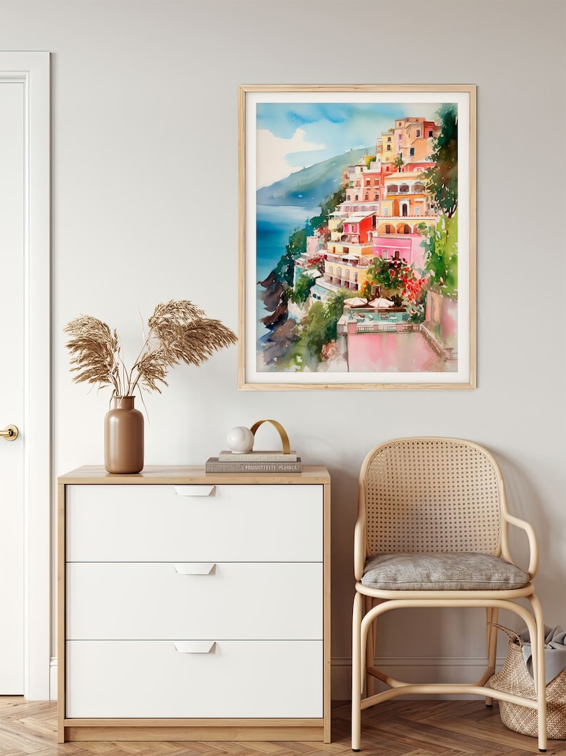 May include: A watercolour painting depicting colourful buildings perched on a cliff overlooking the sea. The buildings are painted in shades of pink, yellow, orange, and red. The sea is a deep blue, and the sky is a light blue with white clouds.