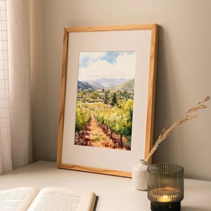 May include: A framed watercolor painting of a vineyard with rolling hills in the background. The painting is in a light wood frame and is sitting on a white table with a book and a candle holder.