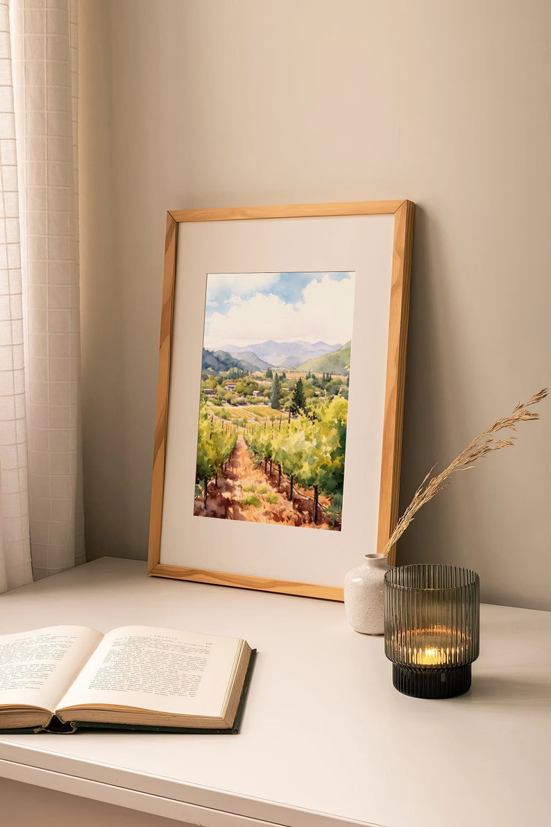 May include: A framed watercolor painting of a vineyard with rolling hills in the background. The painting is in a light wood frame and is sitting on a white table with a book and a candle holder.