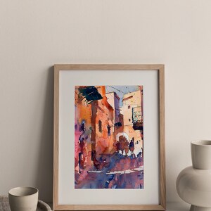 May include: Watercolor painting of a street scene in warm tones, framed in a light wood frame. The artwork depicts buildings with arched doorways and figures, with shades of orange, red, and blue. The painting is displayed on a white surface.