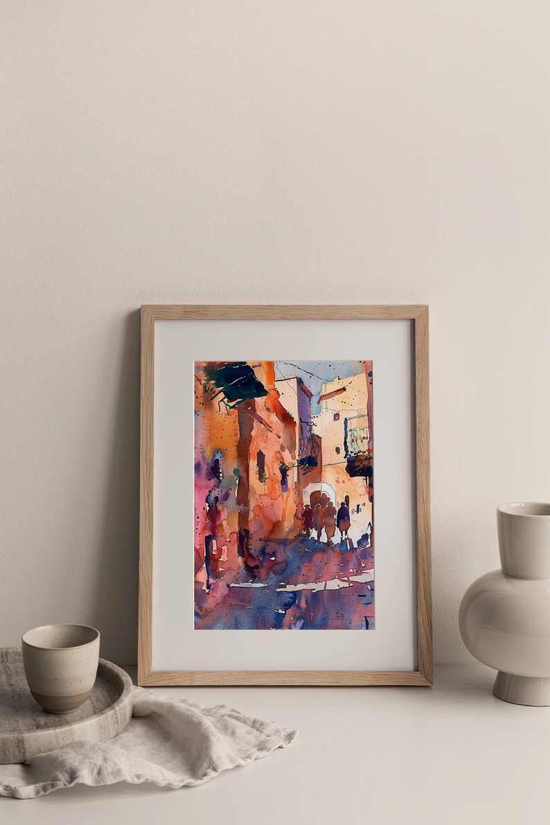 May include: Watercolor painting of a street scene in warm tones, framed in a light wood frame. The artwork depicts buildings with arched doorways and figures, with shades of orange, red, and blue. The painting is displayed on a white surface.
