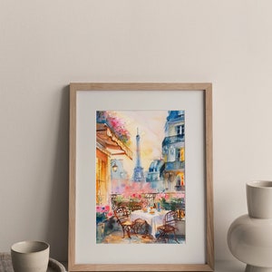 May include: A watercolor painting of a Parisian cafe scene with a view of the Eiffel Tower. The cafe is on a balcony with a table set for two, surrounded by pink flowers. The painting is framed in a light wood frame.