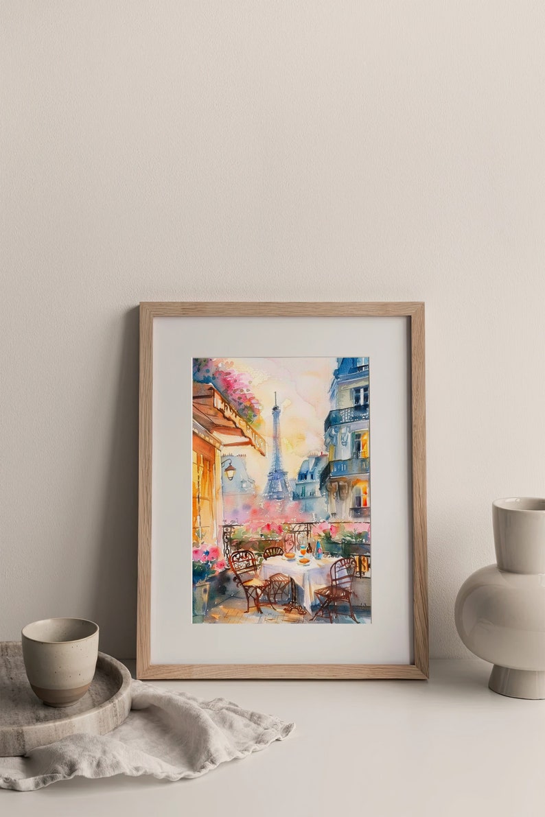 May include: A watercolor painting of a Parisian cafe scene with a view of the Eiffel Tower. The cafe is on a balcony with a table set for two, surrounded by pink flowers. The painting is framed in a light wood frame.