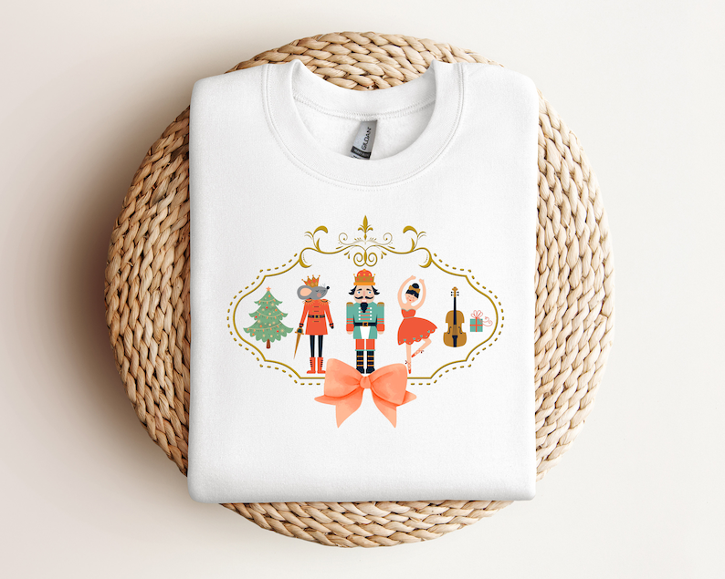 May include: White sweatshirt with a festive Nutcracker design. The design features a Nutcracker soldier, a ballerina, a mouse, a Christmas tree, a violin, and a gift box. The design is framed by a gold border with a bow.