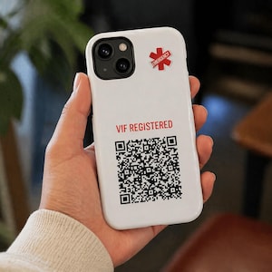 May include: White phone case with a black camera module and a red emergency symbol sticker. The case has the text "VIF REGISTERED" in red, along with a black and white QR code. The phone case is held in a hand.
