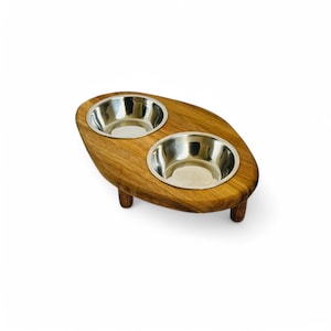 May include: Wooden pet feeder with two stainless steel bowls. The feeder is oval-shaped with a natural wood finish and four short legs. The bowls are set into the wood, creating a raised feeding station for pets.