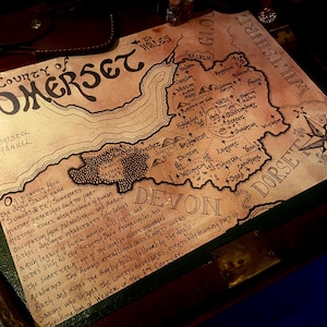 Könnte beinhalten: Eine detaillierte, handgezeichnete Karte der Grafschaft Somerset auf gealtertem Papier. Die Karte zeigt geografische Details, Ortsnamen und eine Windrose. Der Text enthält den Namen der Grafschaft und historische Notizen. Die Karte liegt auf einer dunklen Oberfläche.