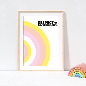 Peut inclure: Une affiche encadrée avec le texte "RESPECT MY PRONOUNS" au-dessus d'un motif arc-en-ciel coloré en jaune, rose et blanc. Un jouet d'empilage arc-en-ciel en bois est à droite de l'affiche encadrée.
