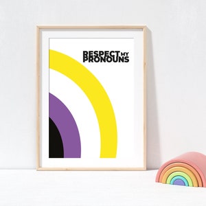 May include: Framed art print with the text "RESPECT MY PRONOUNS" above a non-binary pride flag design. The artwork features a yellow, purple, and black arc on a white background. A wooden rainbow toy sits to the right.