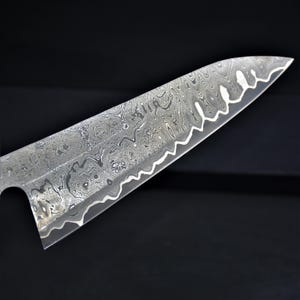May include: Close-up of a stainless steel chef's knife blade with a unique, patterned design. The blade has a wavy edge and a textured surface, showcasing a blend of silver and gray tones. The knife is designed for kitchen use.