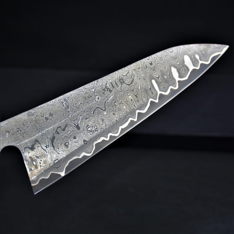 May include: Close-up of a stainless steel chef's knife blade with a unique, patterned design. The blade has a wavy edge and a textured surface, showcasing a blend of silver and gray tones. The knife is designed for kitchen use.