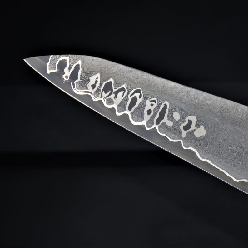 May include: Close-up of a stainless steel chef's knife blade with a unique pattern. The blade has a wavy, marbled design and a decorative edge. The knife is set against a black background.