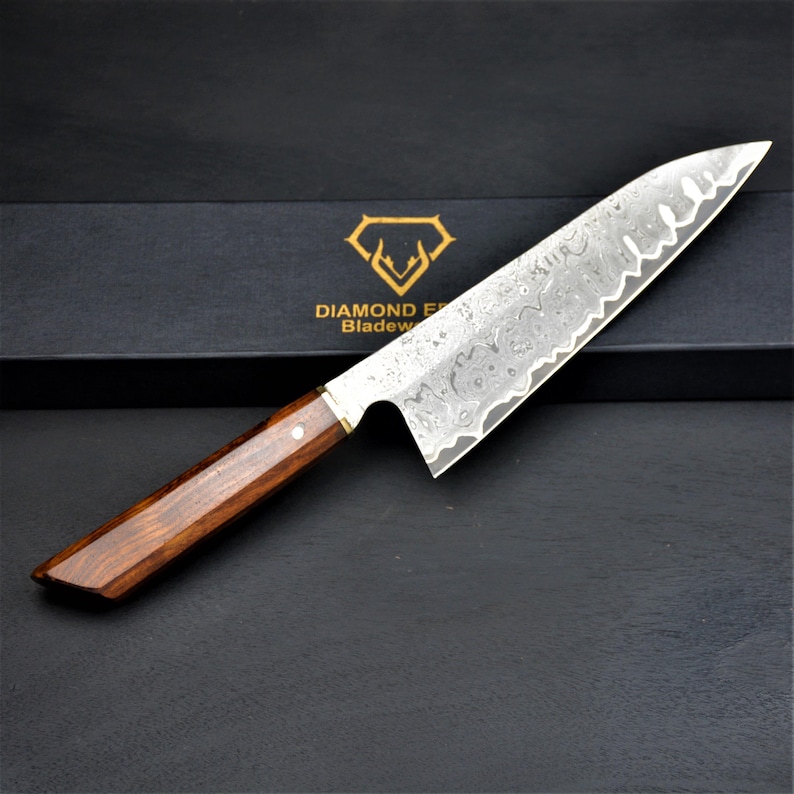 May include: A Damascus steel chef's knife with a wooden handle. The blade has a unique patterned design and a sharp edge. The handle is a dark brown color. The knife is displayed on a black surface.