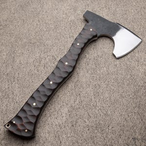 May include: A handcrafted axe with a dark, textured wooden handle and a polished steel blade. The handle features a carved pattern with small, round, brass-colored accents. The axe head is a dark gray color.