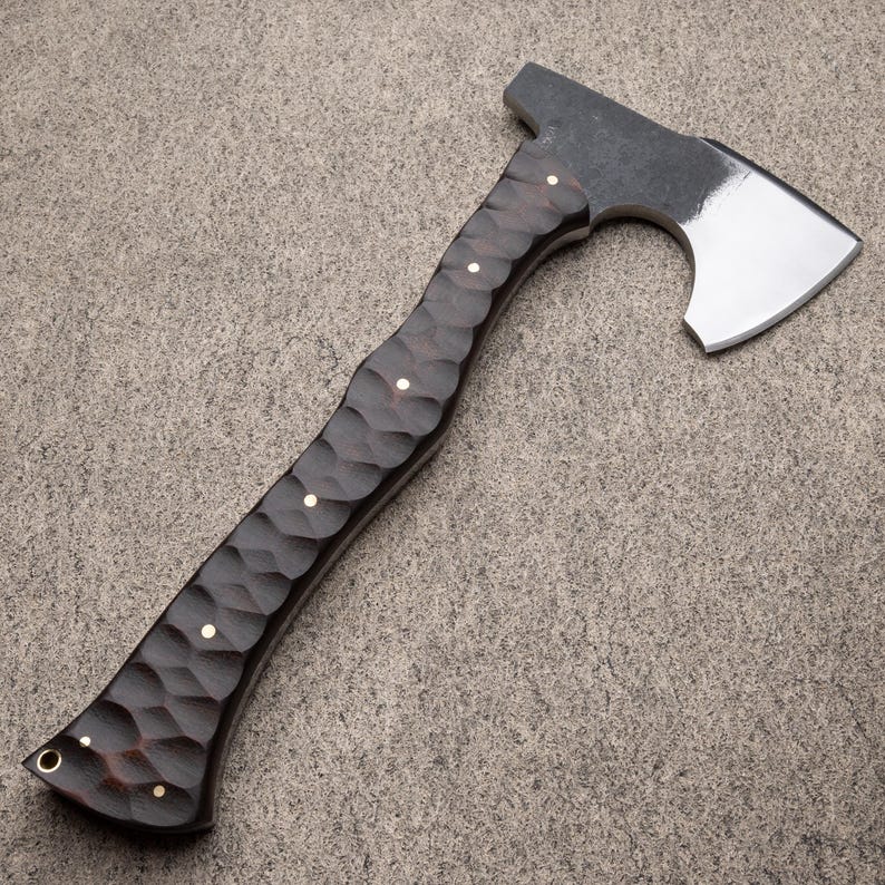 May include: A handcrafted axe with a dark, textured wooden handle and a polished steel blade. The handle features a carved pattern with small, round, brass-colored accents. The axe head is a dark gray color.