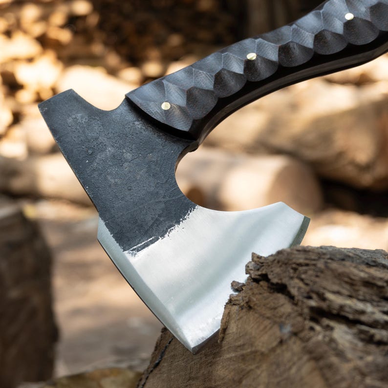 May include: Close-up of a black axe with a silver blade. The handle is dark with a textured pattern and brass rivets. The axe head is resting on a piece of wood, suggesting outdoor or camping use.