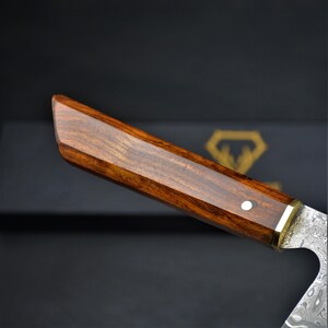 May include: Close-up of a knife handle with a dark wood grain. The handle is rectangular with a brass accent and a single white dot. The blade has a patterned design.