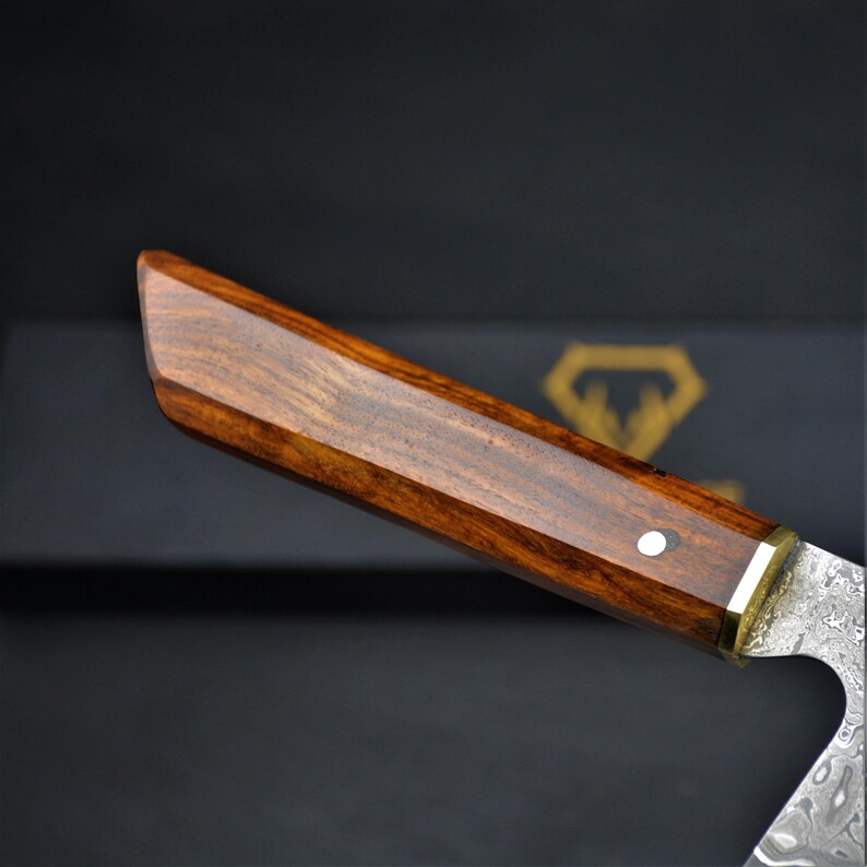 May include: Close-up of a knife handle with a dark wood grain. The handle is rectangular with a brass accent and a single white dot. The blade has a patterned design.