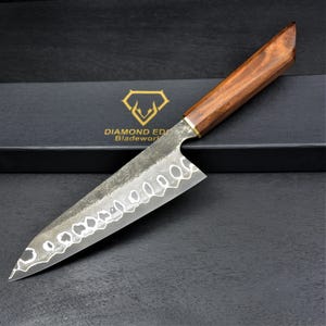 May include: A chef knife with a patterned Damascus steel blade and a polished brown wooden handle. The blade features a distinct wavy metal texture. Behind the knife lies a black box with the gold text DIAMOND EDGE BLADEWORKS and a logo.