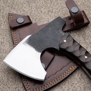 May include: A small, handcrafted axe with a dark, textured blade and a polished, white cutting edge. The handle is dark wood with a carved pattern and brass accents. It rests on a brown leather sheath with decorative stitching and a strap.