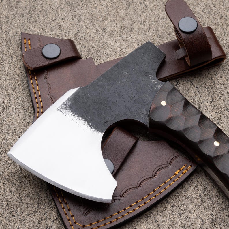 May include: A small, handcrafted axe with a dark, textured blade and a polished, white cutting edge. The handle is dark wood with a carved pattern and brass accents. It rests on a brown leather sheath with decorative stitching and a strap.