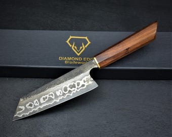 May include: A kitchen knife with a dark, textured Damascus steel blade featuring a wavy pattern and a polished wooden handle with a brass bolster. The knife rests on a black box labeled with the gold text Diamond Edge Bladeworks.