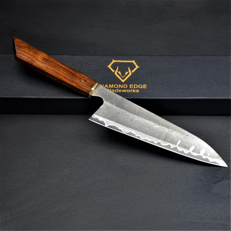 May include: A Damascus steel chef's knife with a wooden handle. The blade has a unique pattern and a sharp edge. The handle is a dark brown color with a brass bolster. The knife is displayed on a black surface with the text "DIAMOND EDGE Tradeworks".