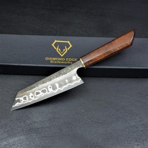 May include: A kitchen knife with a dark, textured metal blade featuring a wavy hamon line and a polished wooden handle. The knife rests on a black box labeled with the gold Diamond Edge Bladeworks logo and text, showcasing a sharp, pointed tip.