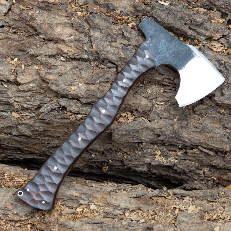 May include: A handcrafted axe with a dark, textured wooden handle and a silver-colored steel blade. The handle features a diamond pattern and is accented with small, white rivets. The axe head has a flat top and a sharp, curved blade.