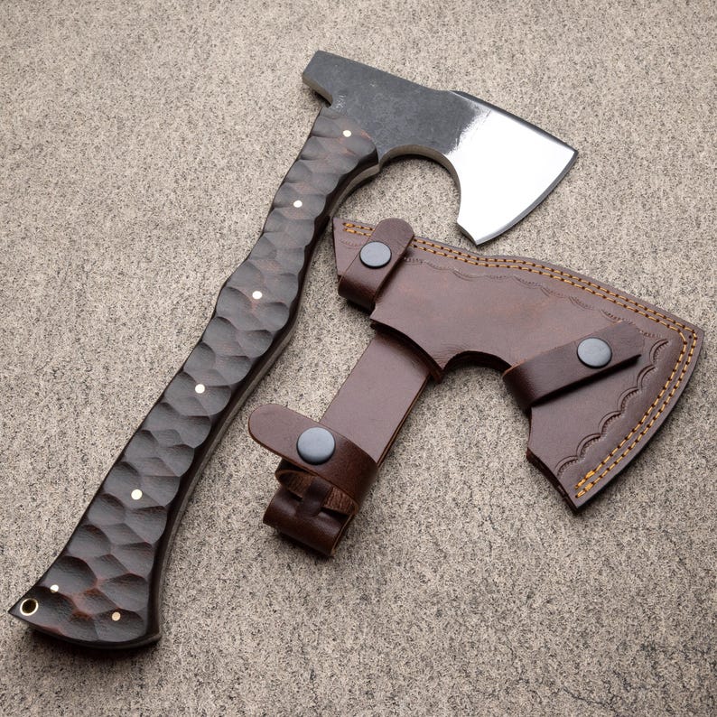 May include: A handcrafted axe with a dark wooden handle and a polished steel blade. The handle features a textured design with brass accents. A brown leather sheath with decorative stitching protects the blade. The axe is designed for outdoor use.