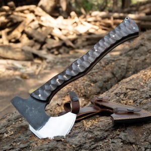 May include: A handcrafted axe with a dark, textured wooden handle and a silver blade. The axe features a leather sheath with a snap closure. The handle has a unique pattern with small, white accents. The axe is resting on a log.