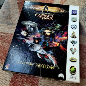 Könnte beinhalten: Spielbox von Star Trek: Starfleet Command Volume II Empires at War. Die Box zeigt einen schwarzen Hintergrund mit einer Weltraumschlacht und einen weißen Rand mit Logos für die verschiedenen Fraktionen im Spiel.