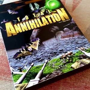 Puede incluir: Caja de videojuego "TOTAL ANNIHILATION" con texto dorado. La portada muestra máquinas de guerra futuristas, aviones y un orbe verde brillante sobre un fondo espacial oscuro. El juego es de Cavedog.
