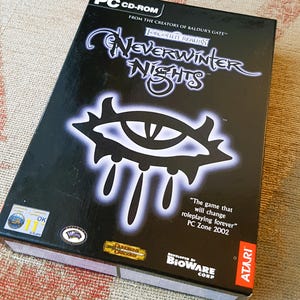 Könnte beinhalten: Eine schwarze Box für das PC-Spiel Neverwinter Nights, mit einem großen, weißen, leuchtenden Auge mit einer schwarzen Pupille. Die Box trägt den Text "Forgotten Realms" und "Das Spiel, das das Rollenspiel für immer verändern wird". Die Box ist mit "PC CD-ROM" beschriftet und hat das Atari-Logo unten rechts.