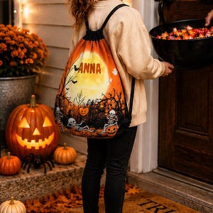Peut inclure: Un sac à dos à cordon avec un imprimé sur le thème d'Halloween représentant une grande lune jaune, des squelettes, des citrouilles et des arbres dénudés. Le nom ANNA est imprimé en lettres grasses sur la lune. Le sac a des bretelles noires.