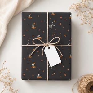 Peut inclure: Un cadeau rectangulaire emballé dans du papier noir avec un motif répétitif de squelettes blancs, de citrouilles orange et d'étoiles orange à quatre branches. Le paquet est noué avec une ficelle fine et porte une étiquette blanche.