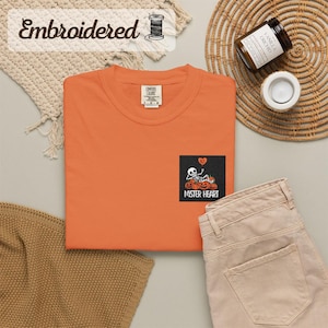 Puede incluir: Una camiseta naranja con un gráfico cuadrado negro en el pecho. El gráfico muestra un esqueleto recostado sobre calabazas con un globo en forma de corazón arriba y el texto MISTER HEART impreso en letras mayúsculas blancas debajo.