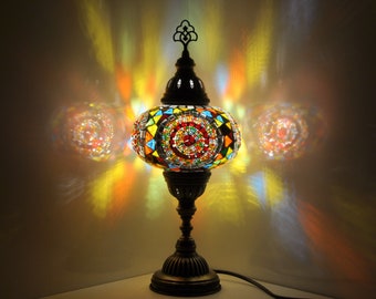Puede incluir: Una lámpara de mesa de vidrio con mosaico de colores con una base redonda y un remate decorativo. La lámpara está encendida y proyecta un brillo cálido.